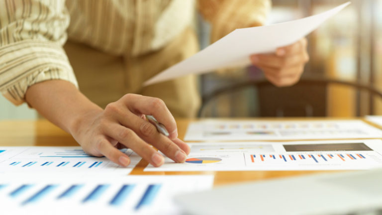close-up view of a businessperson's hand working with paperwork and a laptop on a desk